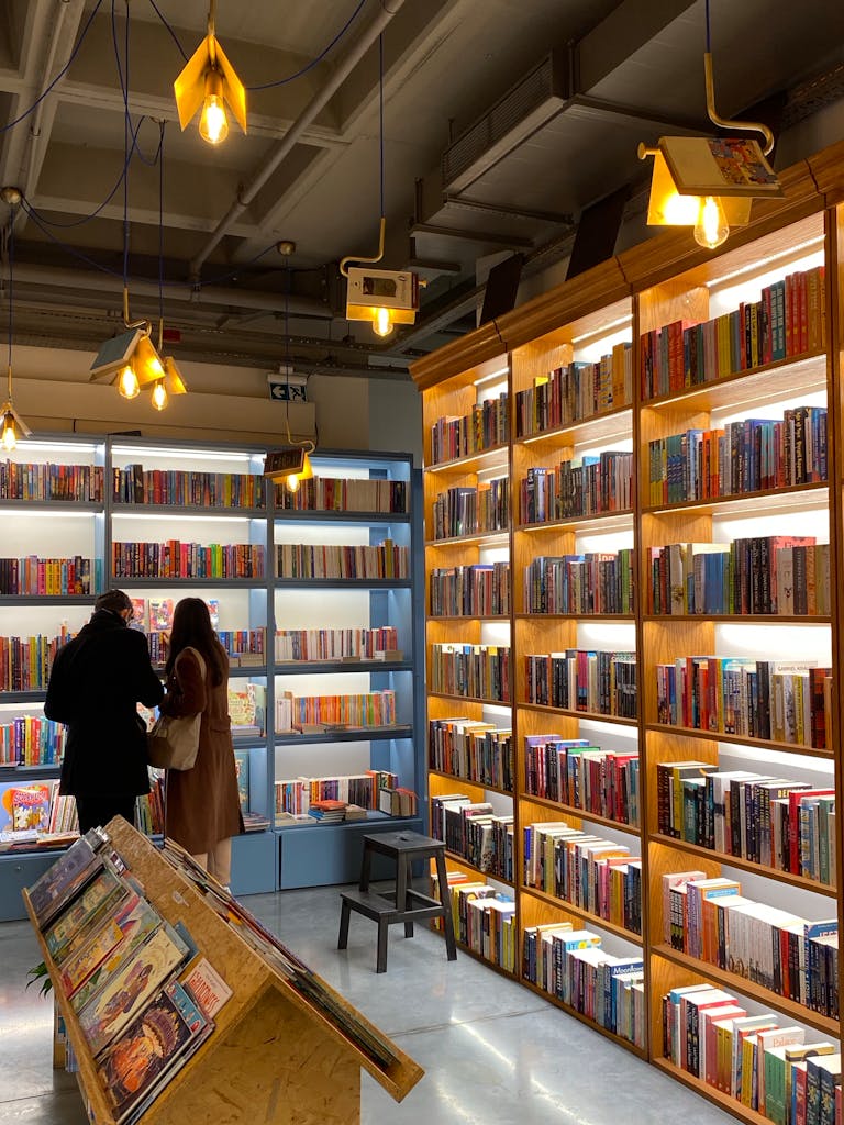 Warmly lit bookstore interior with two customers browsing shelves stocked with books.