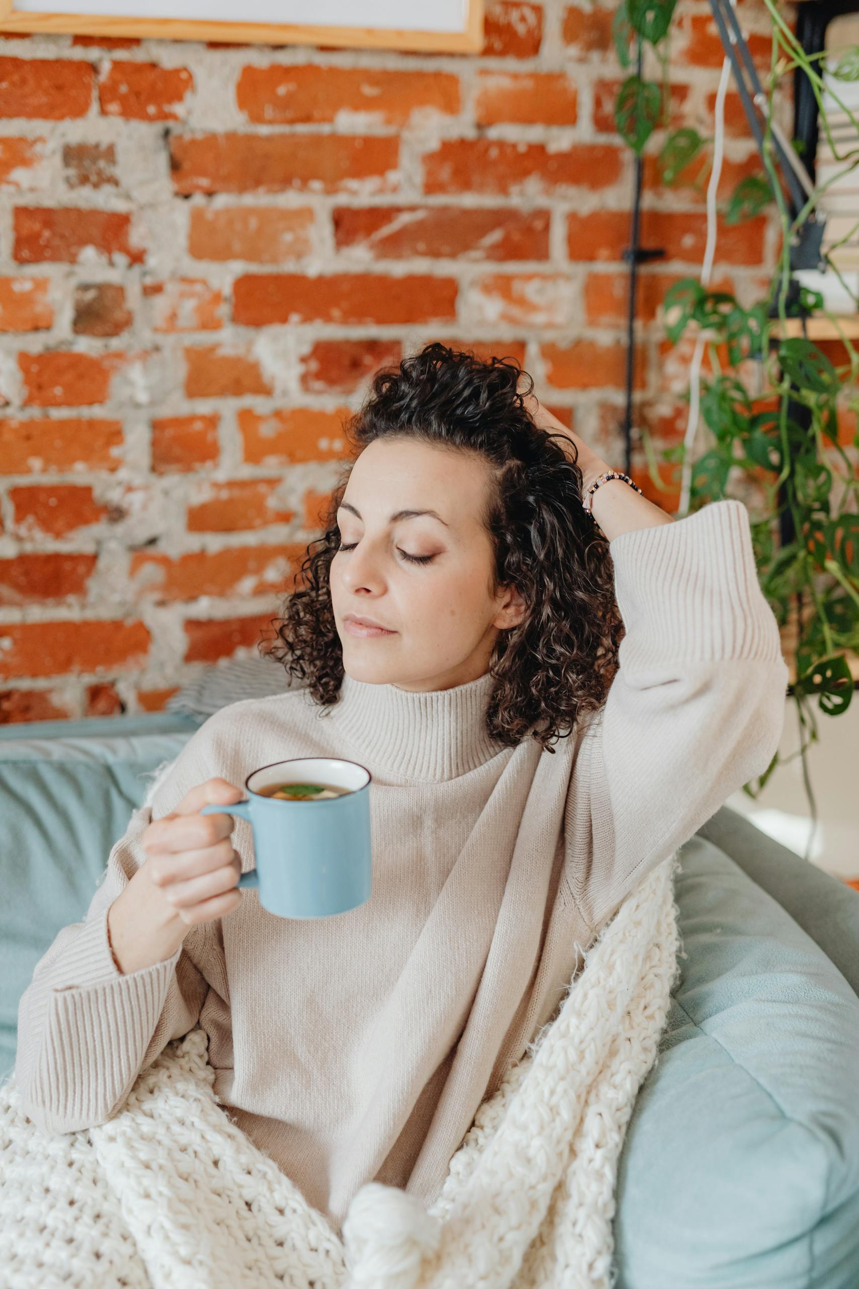 Woman with eyes closed holding a tea cup, relaxing against a brick wall indoors.