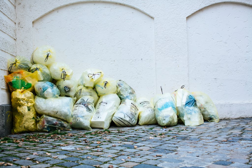 Yellow garbage bags piled against a white wall on a cobblestone street corner outdoors.