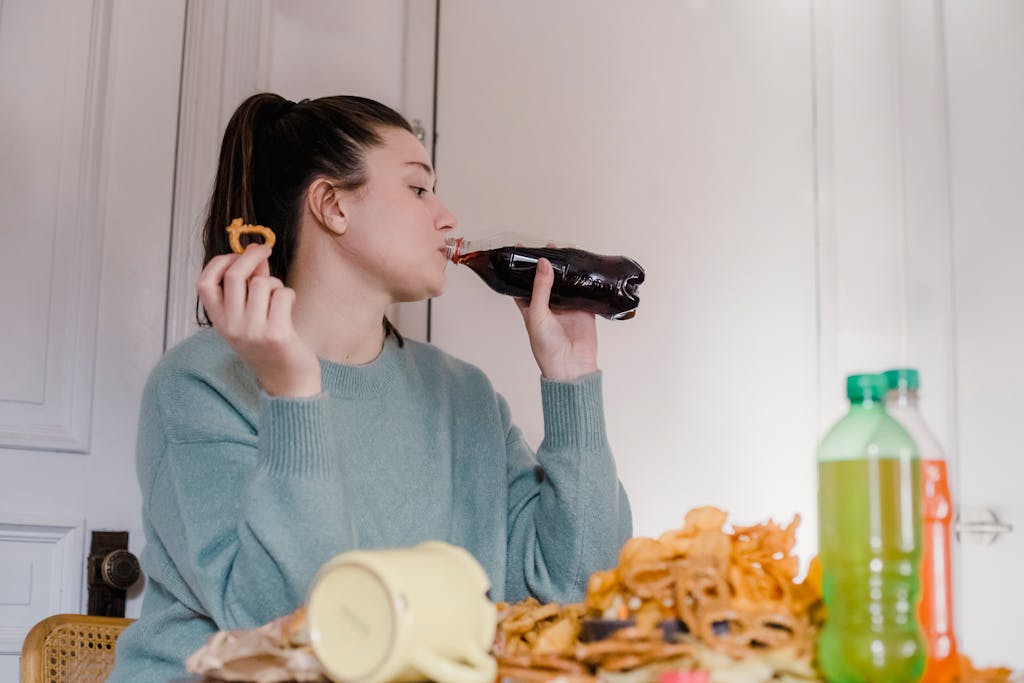 Young female drinking sweet tasty lemonade from plastic bottle at table with different snacks