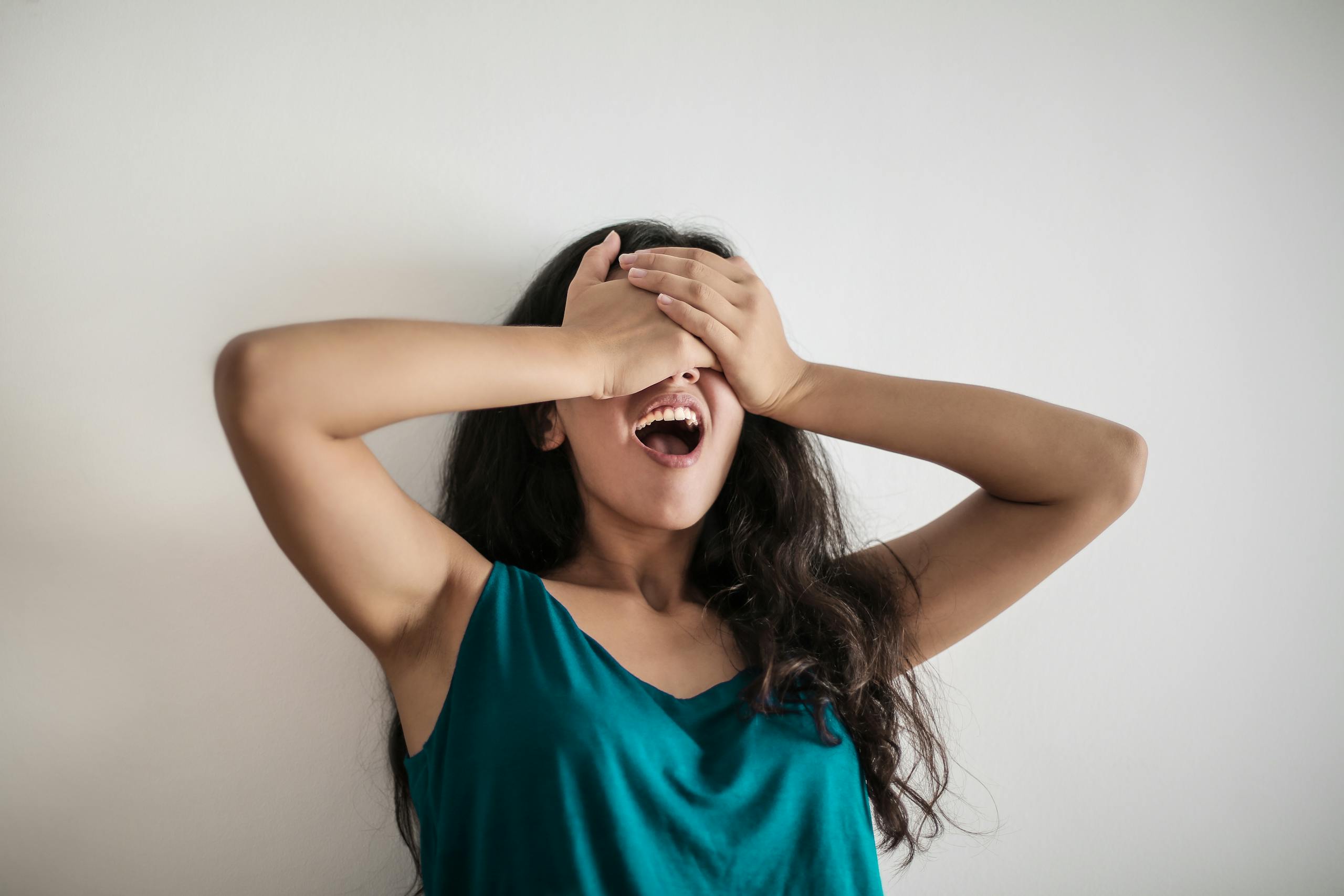 Young long haired female standing with opened mouth and covering eyes with hands in surprise against white background