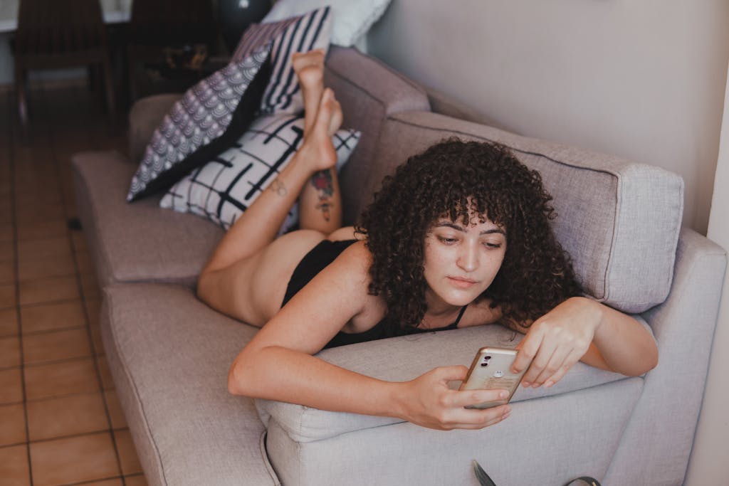 Young woman with curly hair relaxing on a couch, using her smartphone indoors.