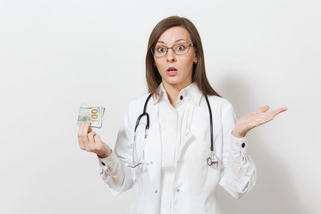 Shocked young doctor woman with stethoscope, glasses isolated on white background. Female doctor in medical gown holding cash money banknotes, pack of dollars. Healthcare personnel, medicine concept