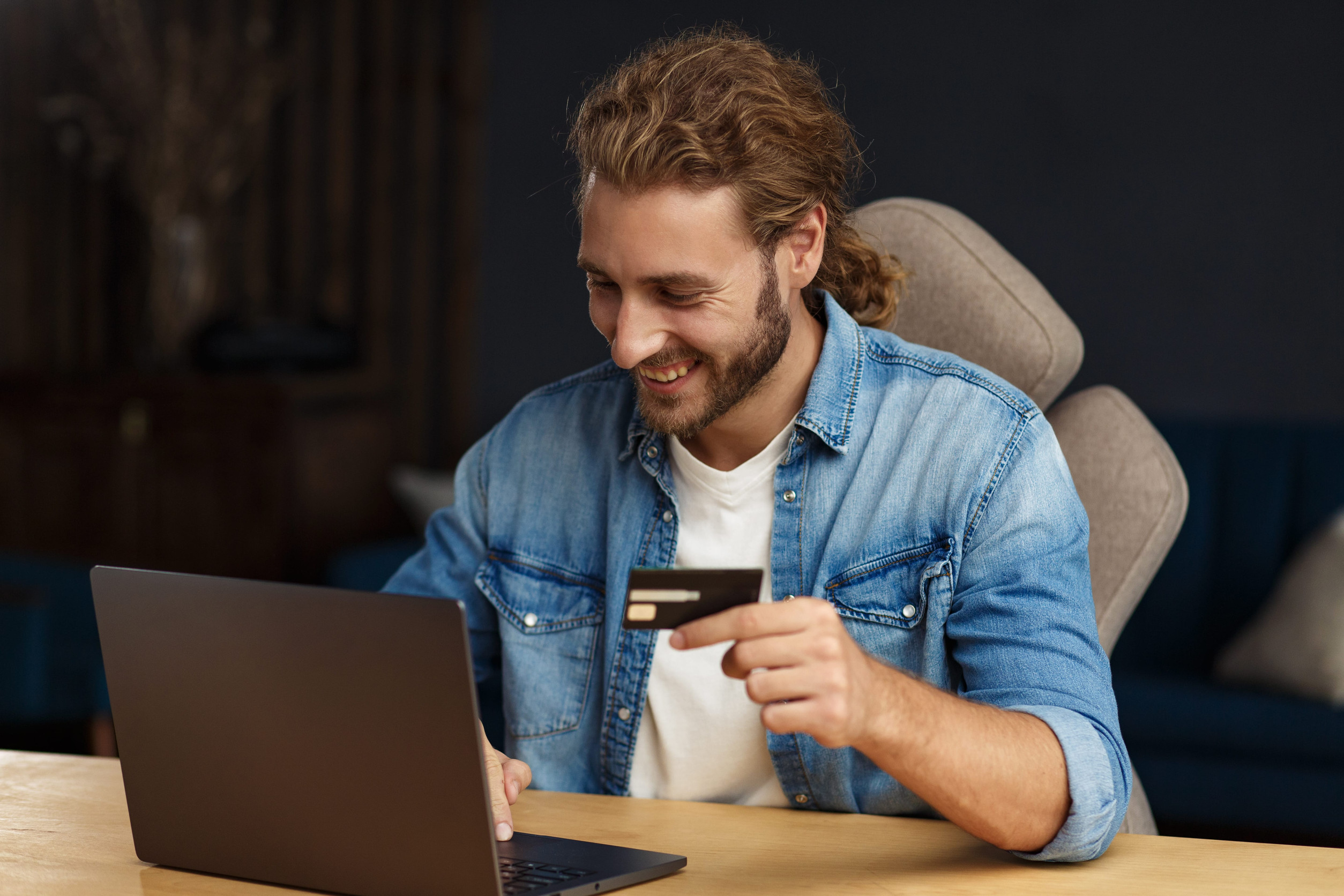 Young handsome curly smiling man with long hair in home office holding credit card and using laptop. Online shopping, online payments. Booking tickets online. Modern technologies concept.