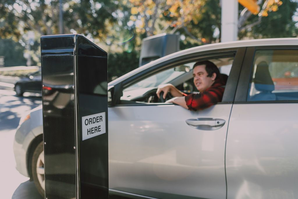 A man in a car orders at a drive-thru window, surrounded by outdoor scenery.