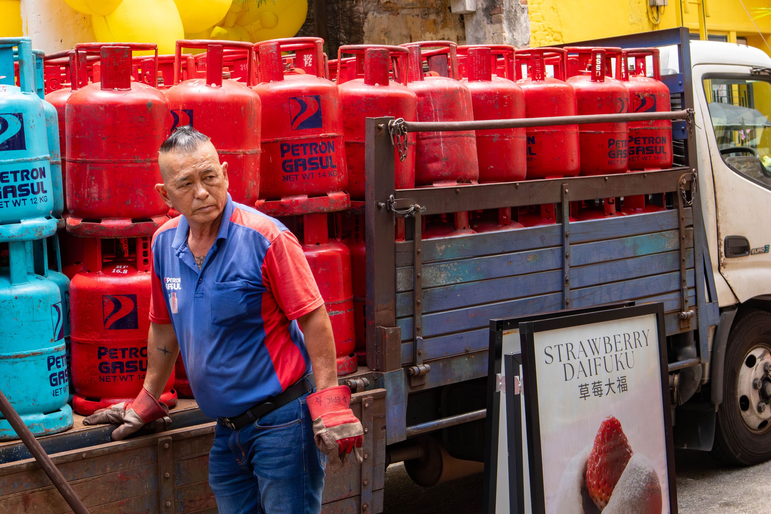 A man in uniform loads red and blue gas cylinders onto a truck in an urban setting.