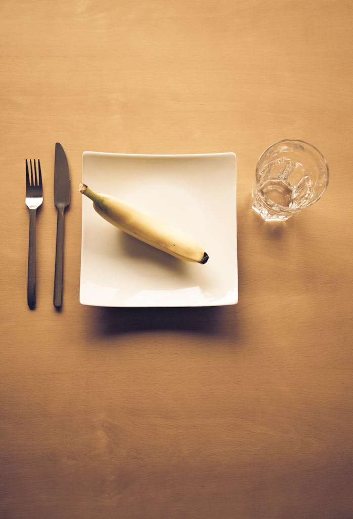 A minimalist dining arrangement featuring a banana on a square plate, with cutlery and a glass.