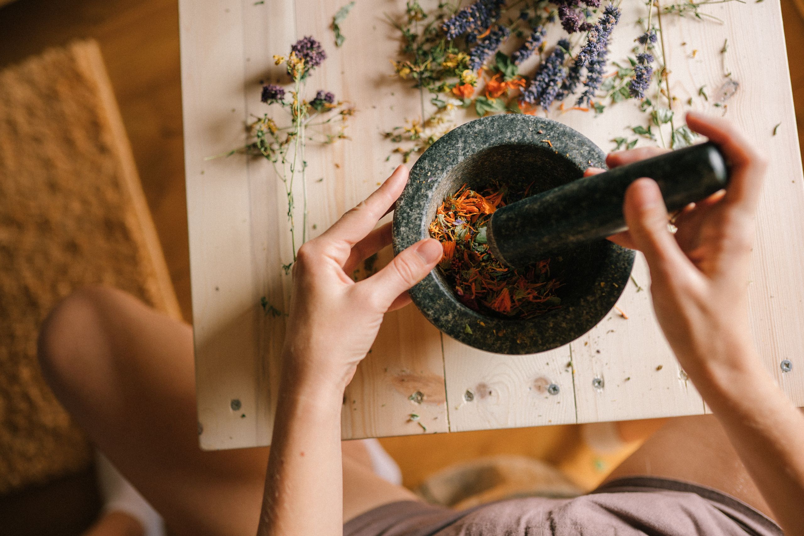 A person grinding colorful dried herbs and flowers in a stone mortar and pestle.