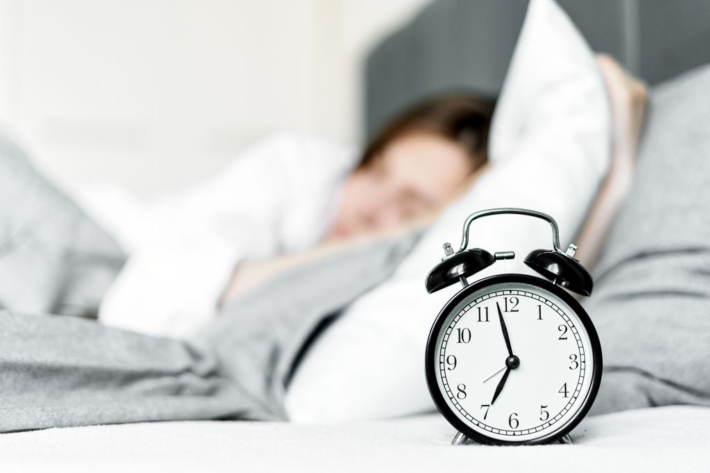 A retro alarm clock sits by a sleeping woman in a cozy bedroom setting.