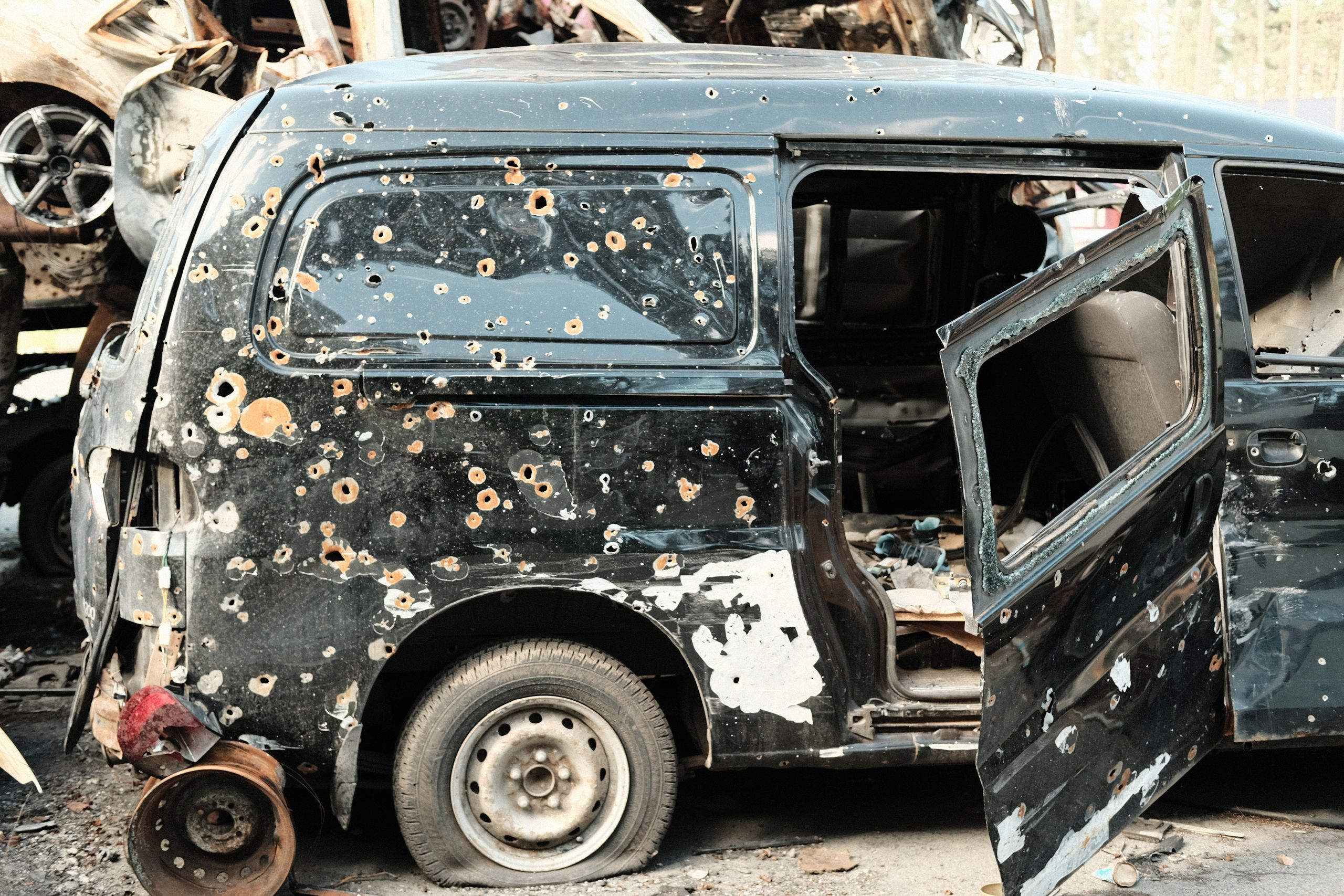 A side view of an abandoned, damaged black van with bullet holes parked in a junkyard.