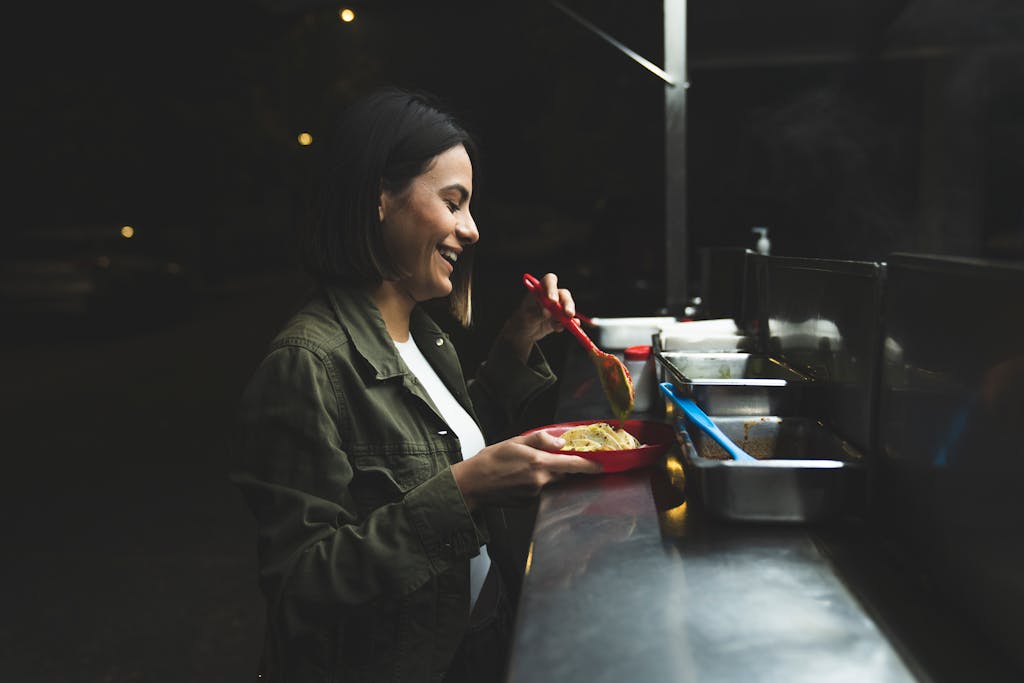 A woman smiling while adding sauce to her tacos at a street stand at night.