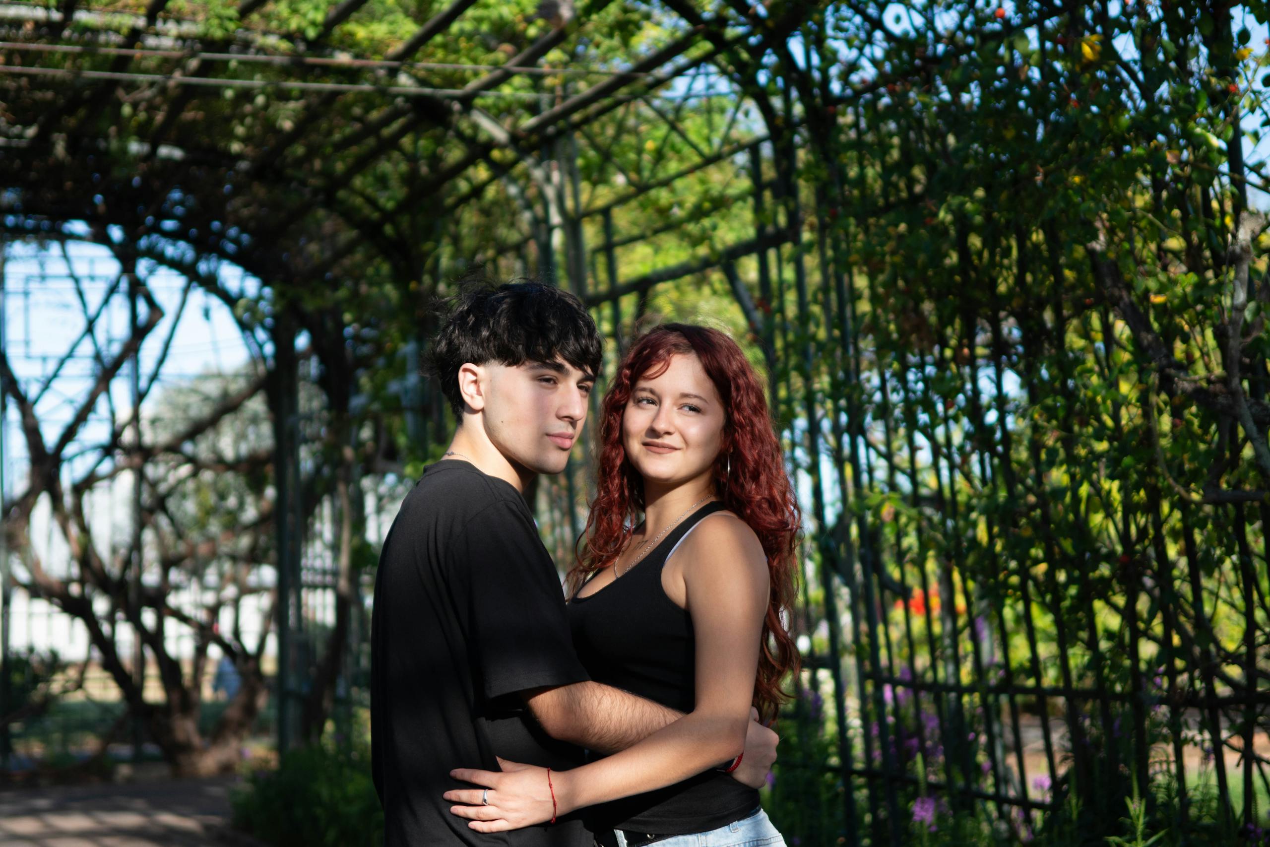 A young couple embraces under a leafy garden trellis on a sunny day, surrounded by nature.