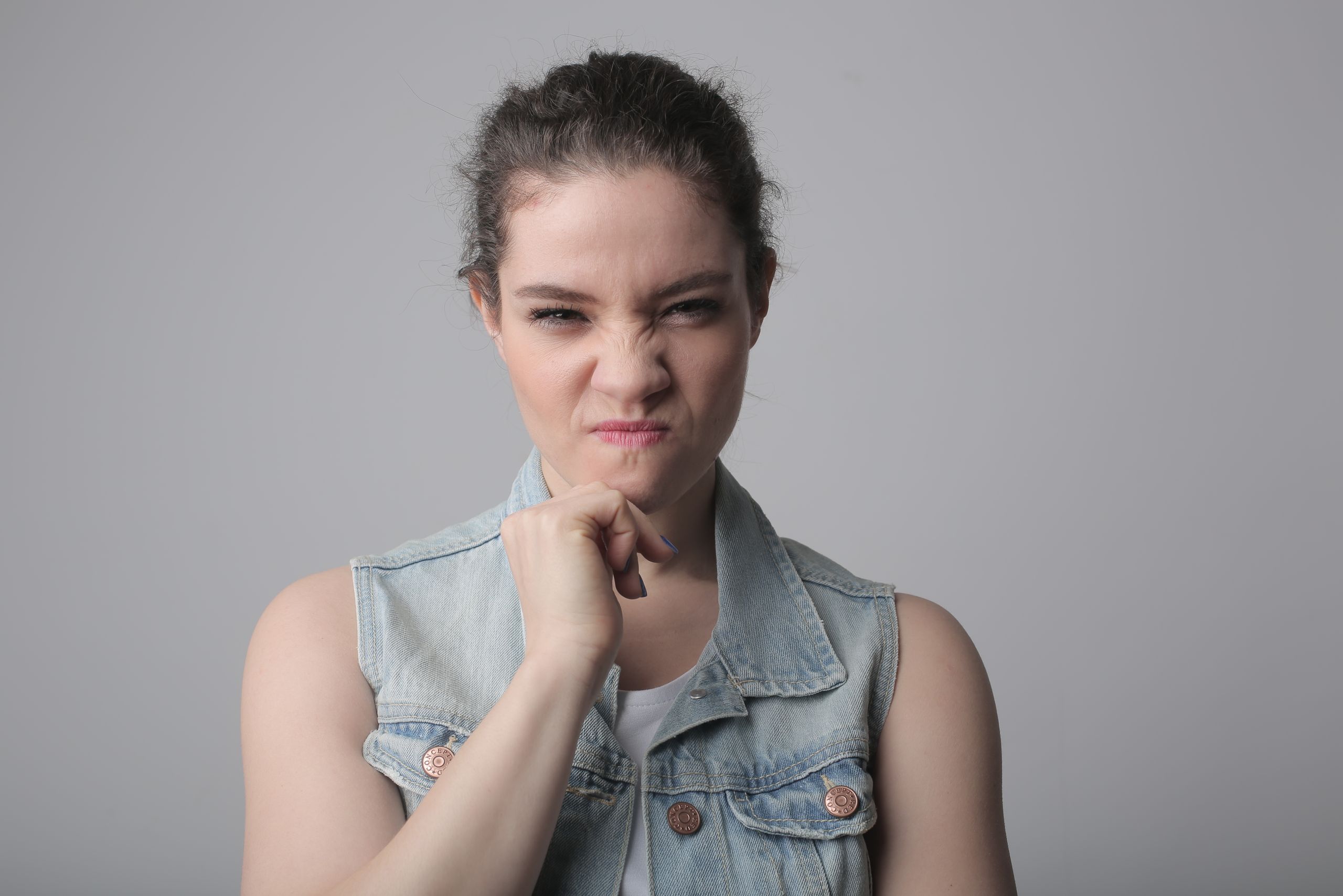 A young woman in a denim vest makes a playful, scrunching face against a neutral background.