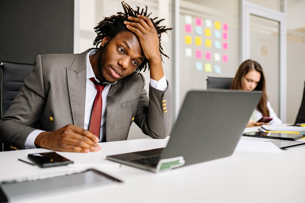 African American man looking stressed in office, with laptop and smartphone on desk.