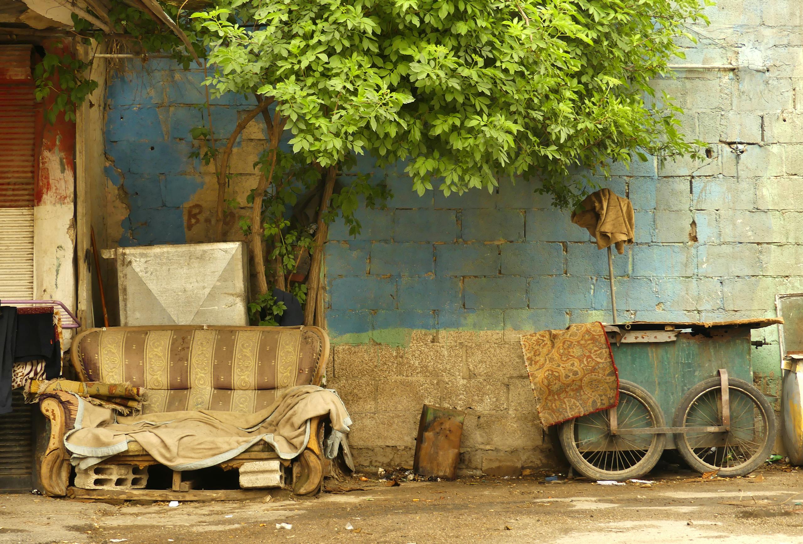An urban street scene in Damascus featuring an abandoned couch under a tree against a blue wall.