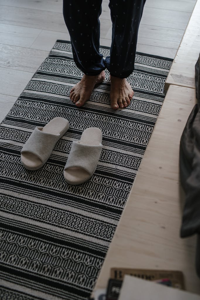 Barefoot person standing beside slippers on a patterned rug indoors.