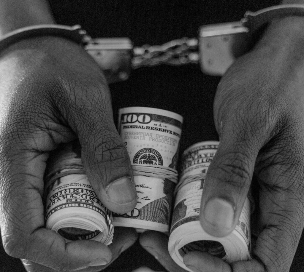 Black and white photo of handcuffed hands holding rolls of US dollar bills.