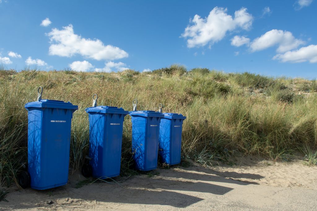 Blue trash bins line up near sandy dunes on a sunny day at Wassenaar beach.
