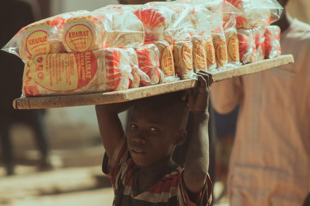 Child street vendor carrying multiple loaves of Khairat butter bread on a wooden tray.