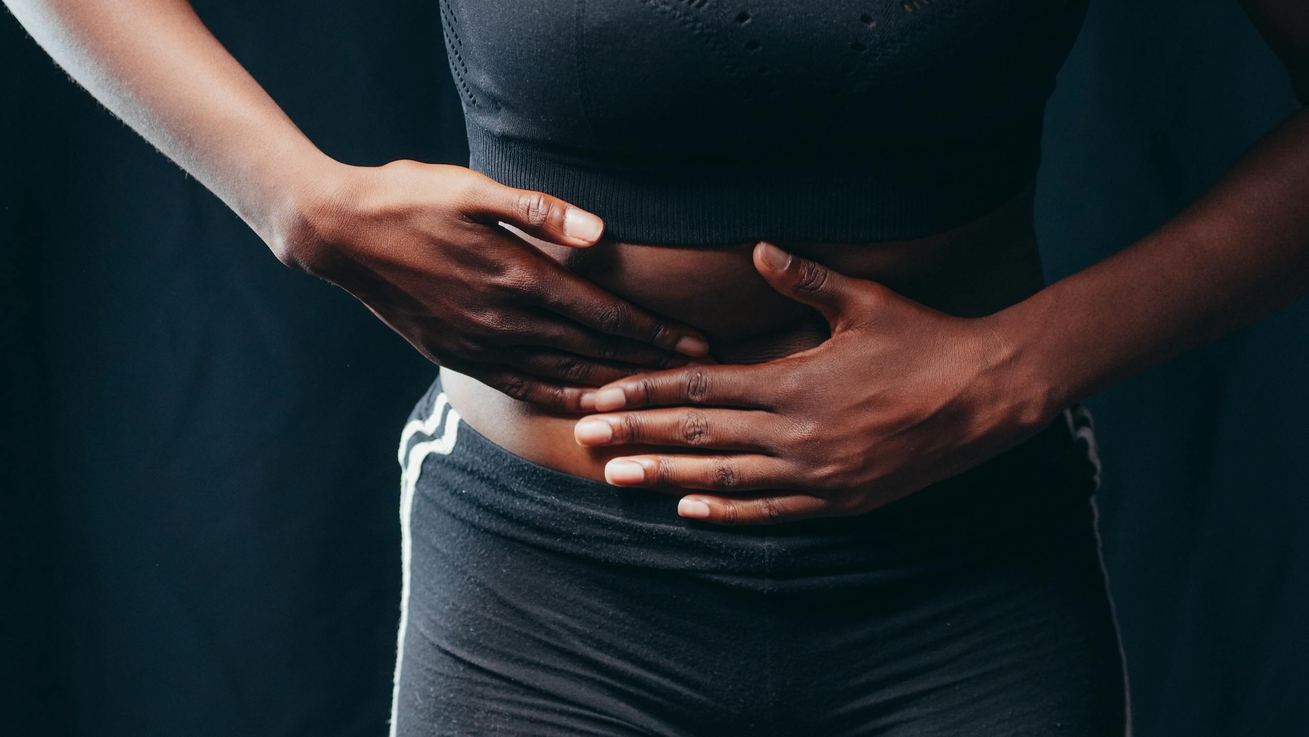 Close-up of a person pressing hands on abdomen, indicating discomfort or pain, against a black background. kidney