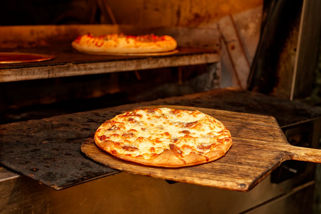 Close-up of cheesy pizza on a wooden peel being baked in a hot oven.