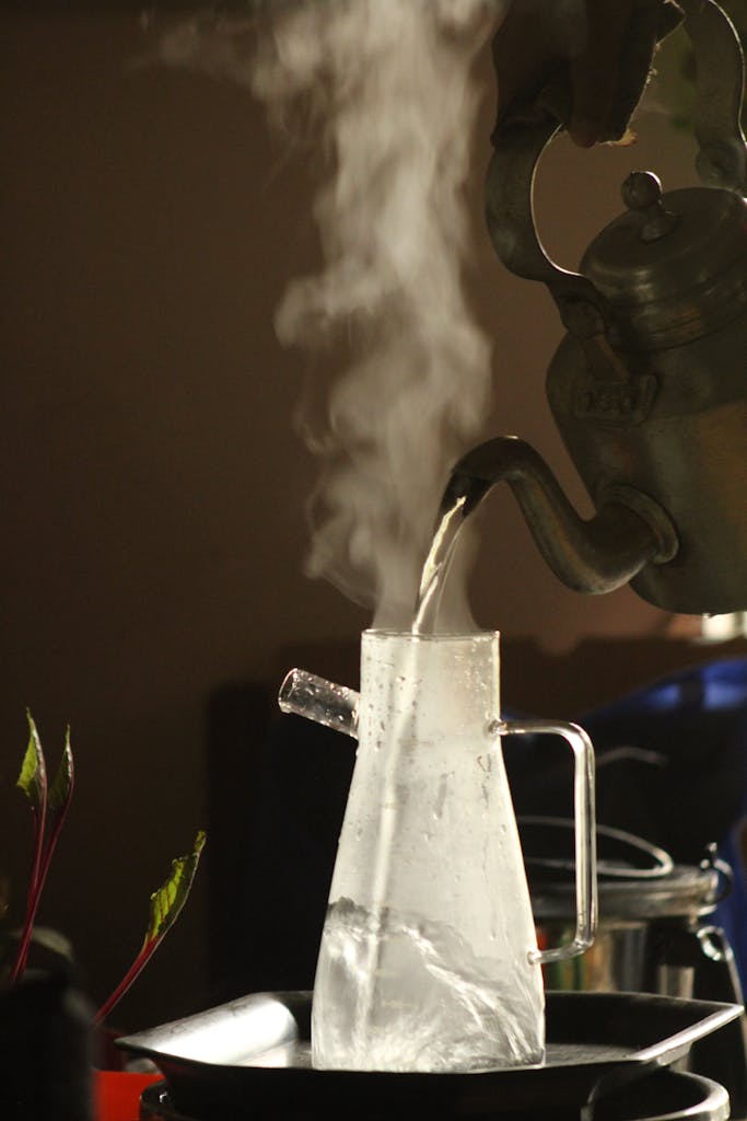 Close-up of steaming hot water being poured from a kettle into a glass pitcher indoors.