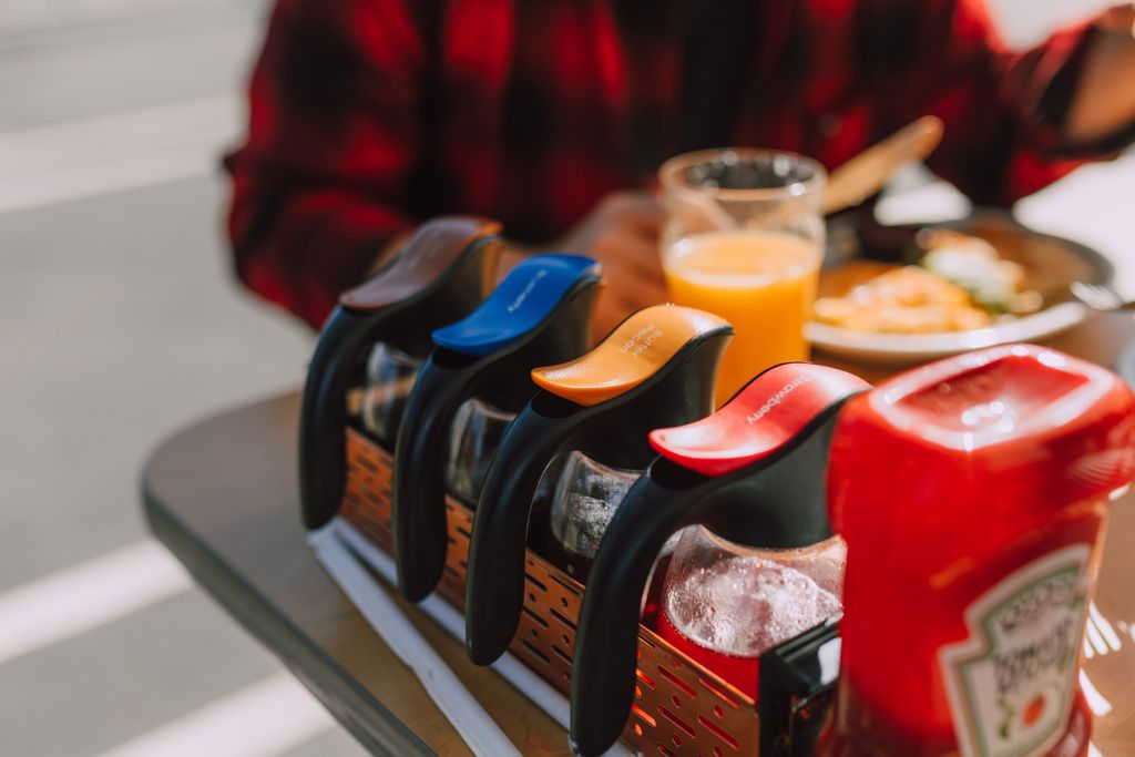 Close-up of syrup dispensers, ketchup, and orange juice on a breakfast table.