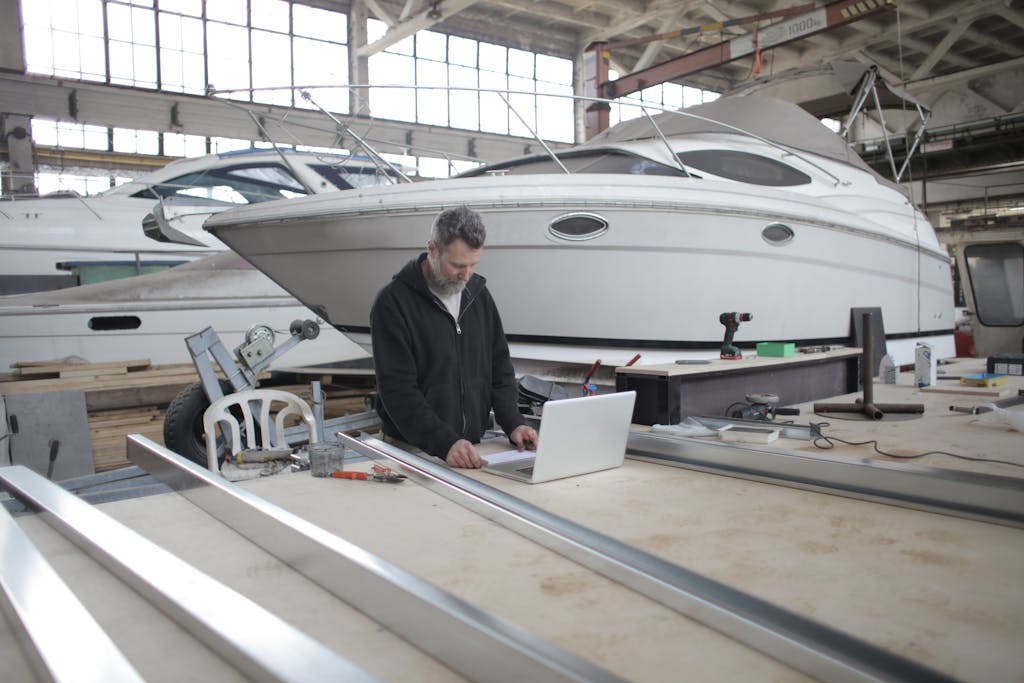 Concentrated adult bearded worker in casual clothes browsing laptop while working with metal details near yacht in garage