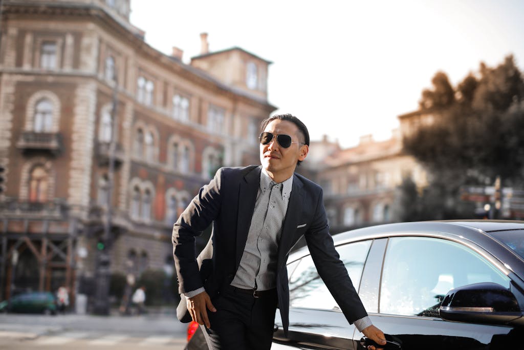 Confident businessman in sunglasses standing by car in city street.