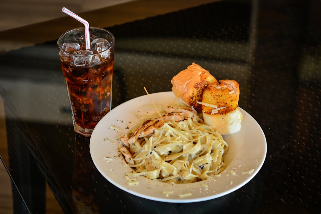 Delicious Italian creamy pasta served with grilled bread and a refreshing cold drink on a glass table.
