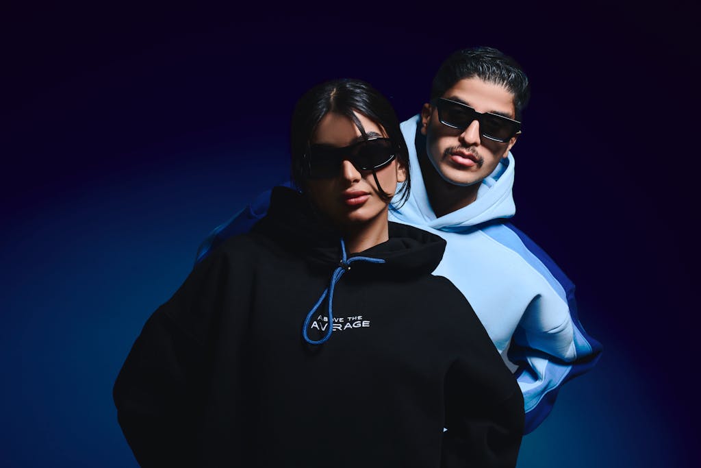 Fashionable couple wearing hoodies and sunglasses, posing confidently under studio lights.