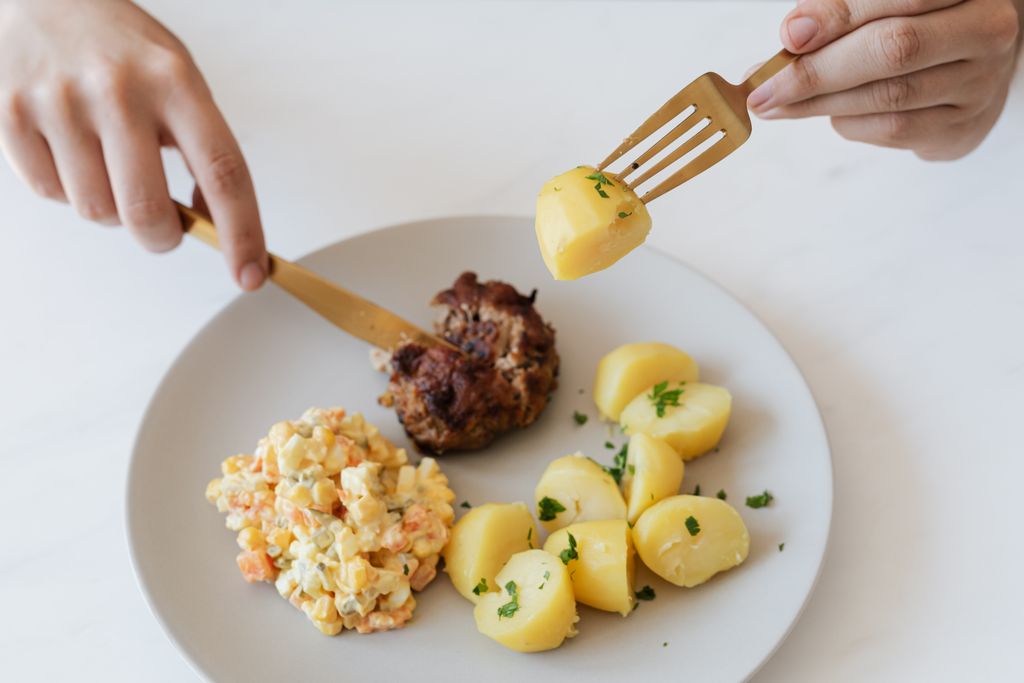 From above of crop anonymous person with cutleries eating delicious fried cutlet and hot boiled potatoes with parsley garnished with nutritious Russian salad