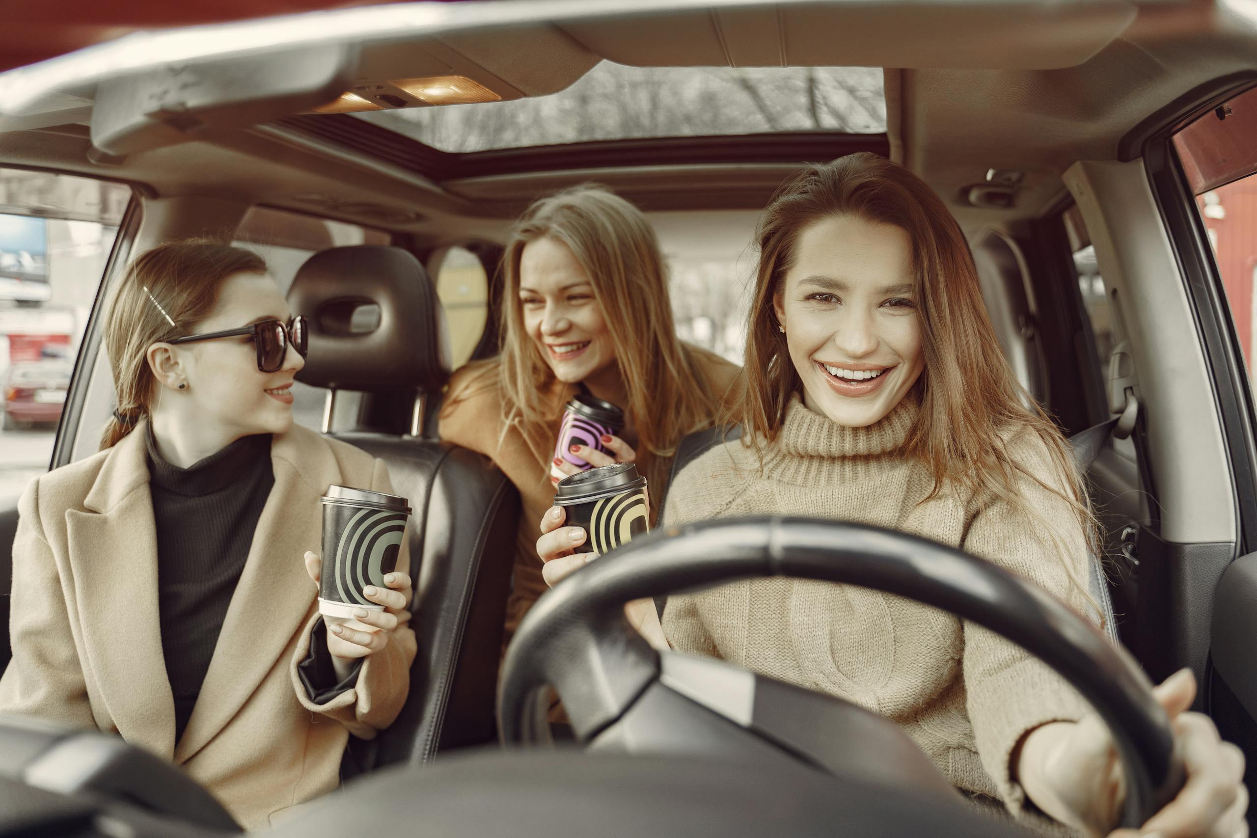 Group of female friends in casual outfit talking in automobile with coffee to go while driving around modern city streets