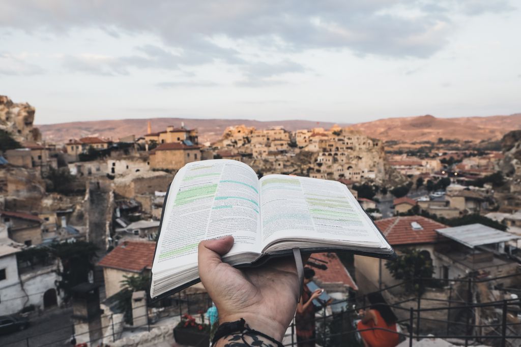 Hand holding an open Bible with a scenic view of ancient Cappadocia buildings in the background.