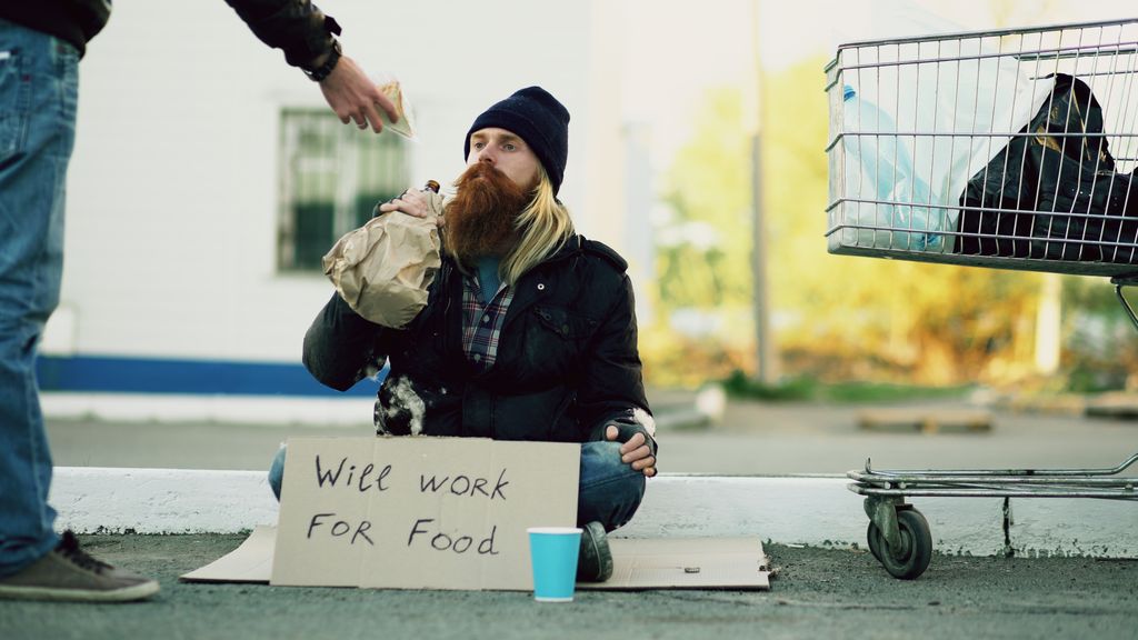 Homeless individual with 'Will Work For Food' sign receives help outdoors.