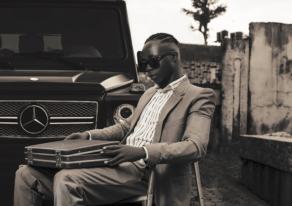 Man in sunglasses sitting outdoors with suitcase near Mercedes-Benz car.