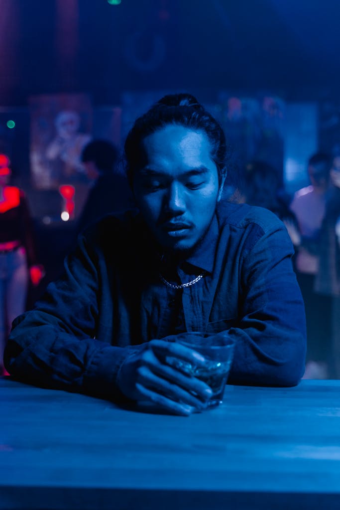 Man sitting alone at a bar holding a drink, surrounded by nightlife ambiance in a dimly lit room.