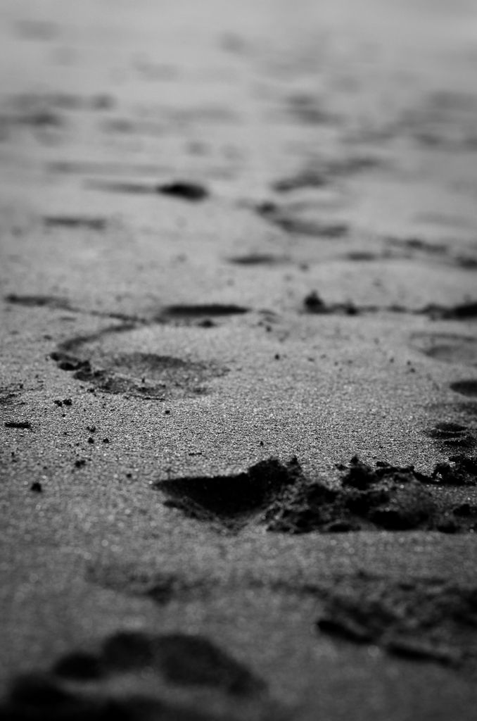 Moody black and white photo of footprints trailing across a black sand beach.