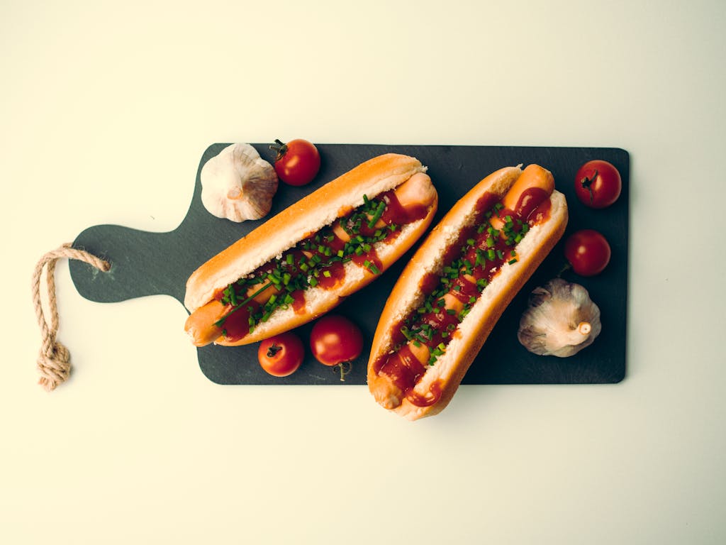 Top view of two delicious hotdogs with tomatoes and garlic on a dark chopping board.