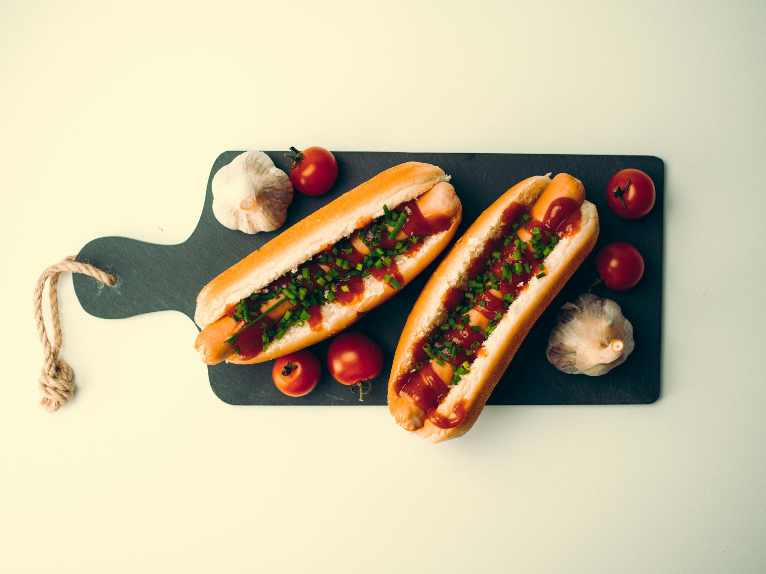 Top view of two delicious hotdogs with tomatoes and garlic on a dark chopping board.