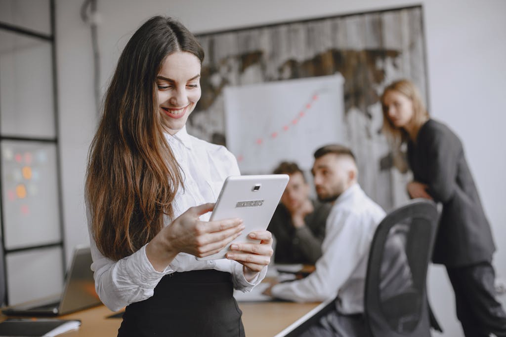 Young woman in a professional office setting browsing a tablet with colleagues in the background.